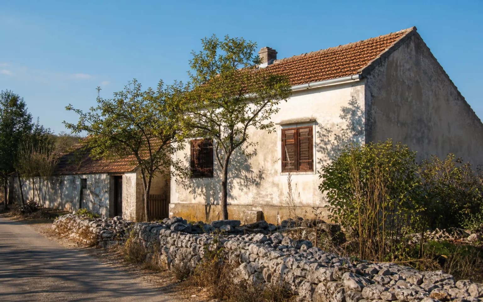 AI-generated illustrative exterior of a modest Dalmatian home with a stone wall, shutters and natural greenery in the Krupa setting.