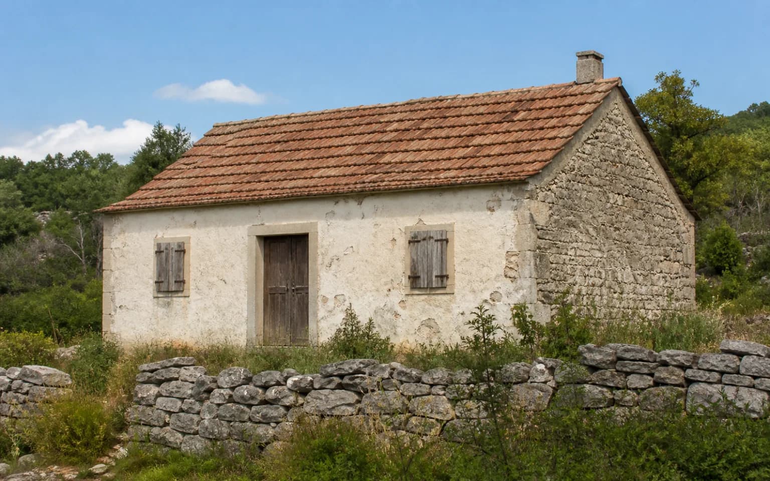 AI ilustrativni eksterijer skromne dalmatinske kuće s istrošenim pročeljem, drvenim škurama i niskim kamenim zidom u ambijentu Zelengrada.