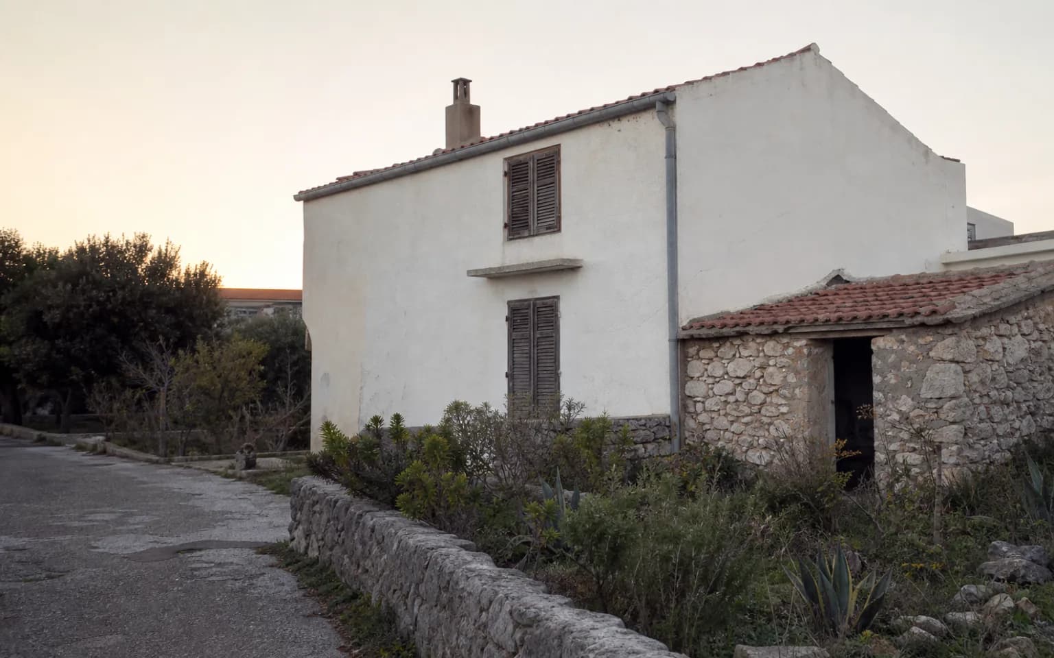 AI-generated illustrative exterior of a modest island home with a pale facade, shutters and a stone annex in the Smokvica, Pag setting.