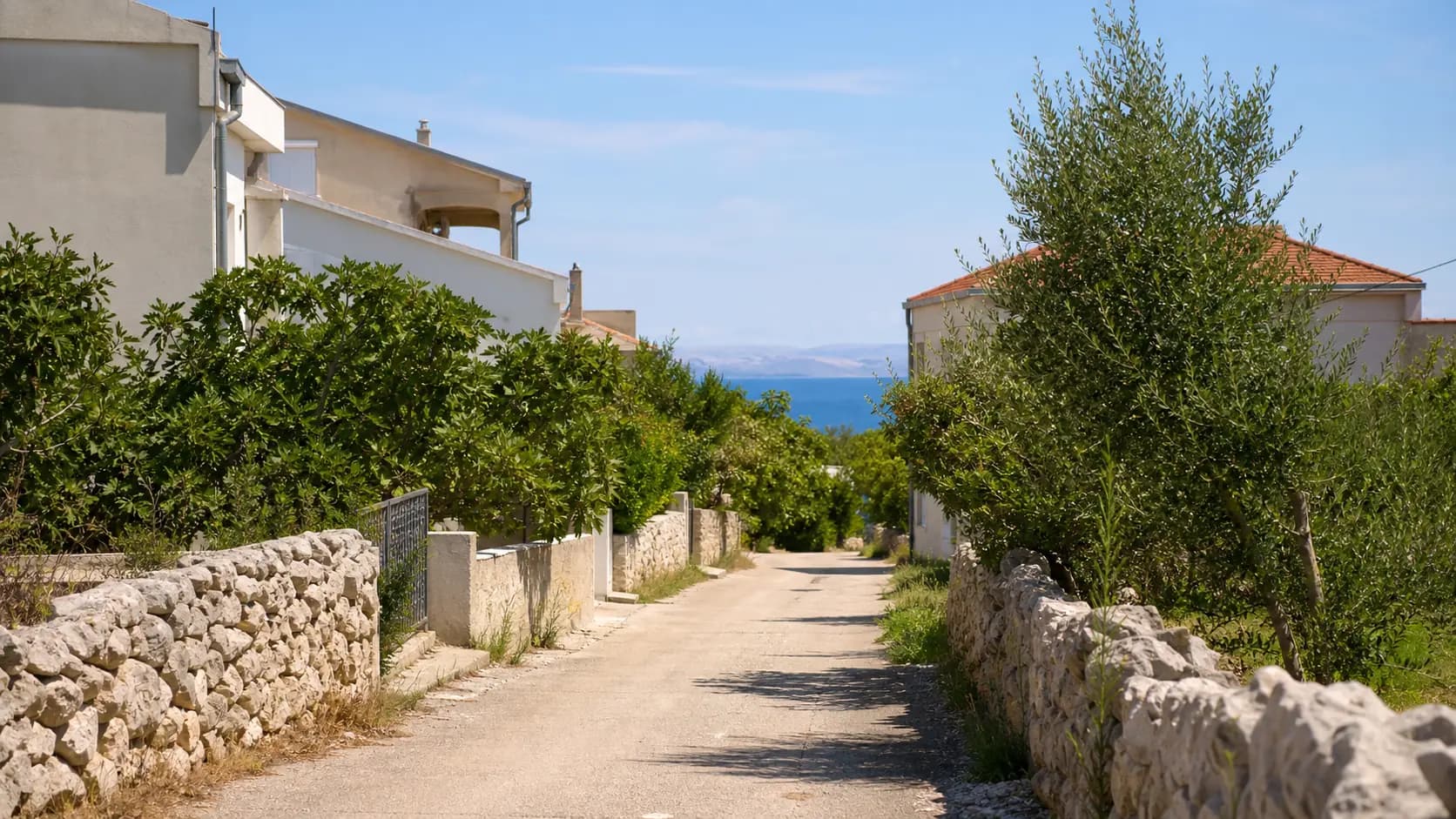 Eine ruhige Straße mit Steinmauern, niedrigen Häusern und fernem Meer in Kolanjski Gajac.
